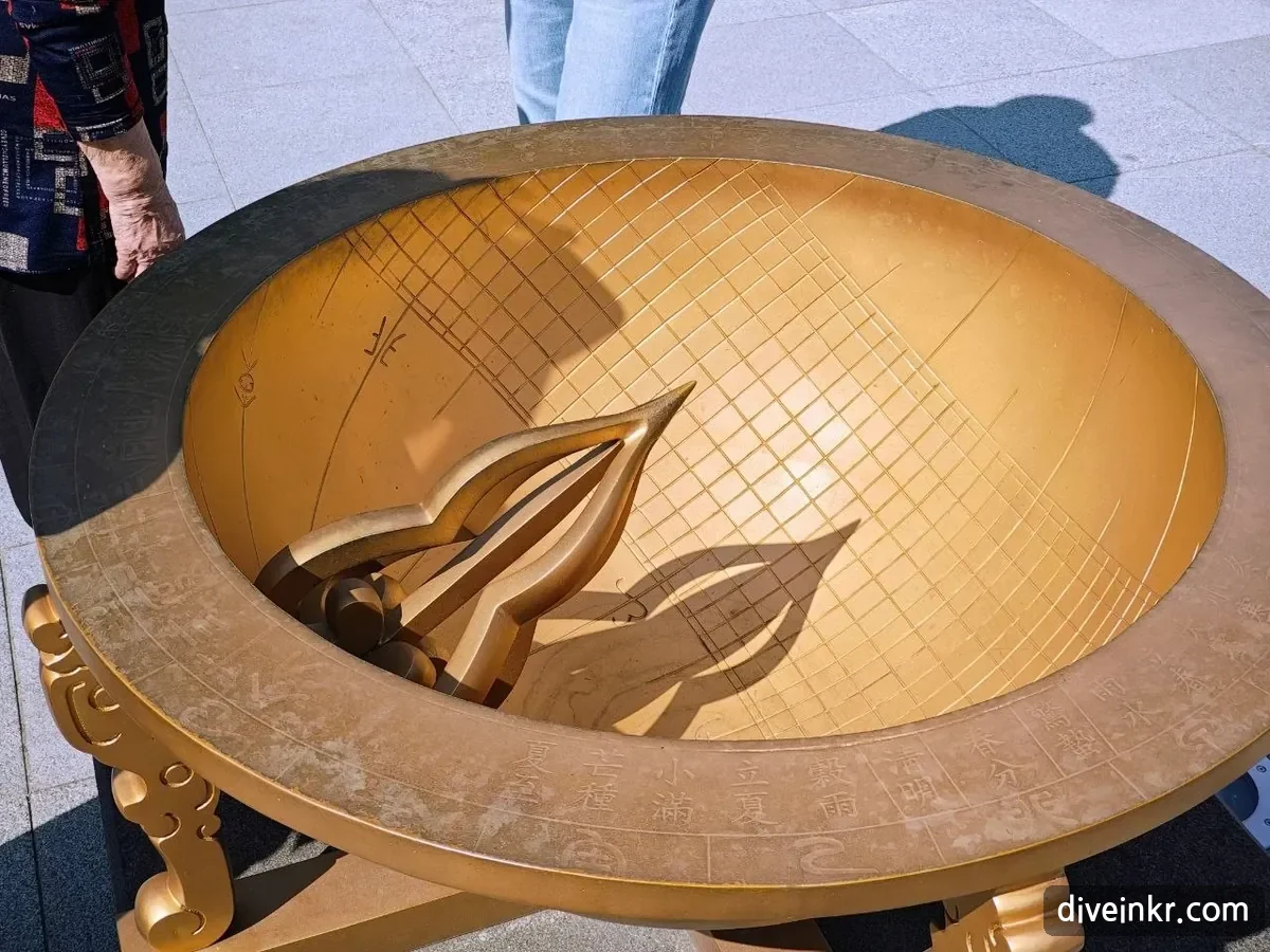 A golden copy of the Joseon hemispherical sundial: a bowl-shaped dial with a grid of hour and season lines, a pointed gnomon casting a shadow, and Chinese characters for the solar terms such as 夏至, 芒種, 小滿, 立夏, 穀雨, 淸明 and 春分 around the rim