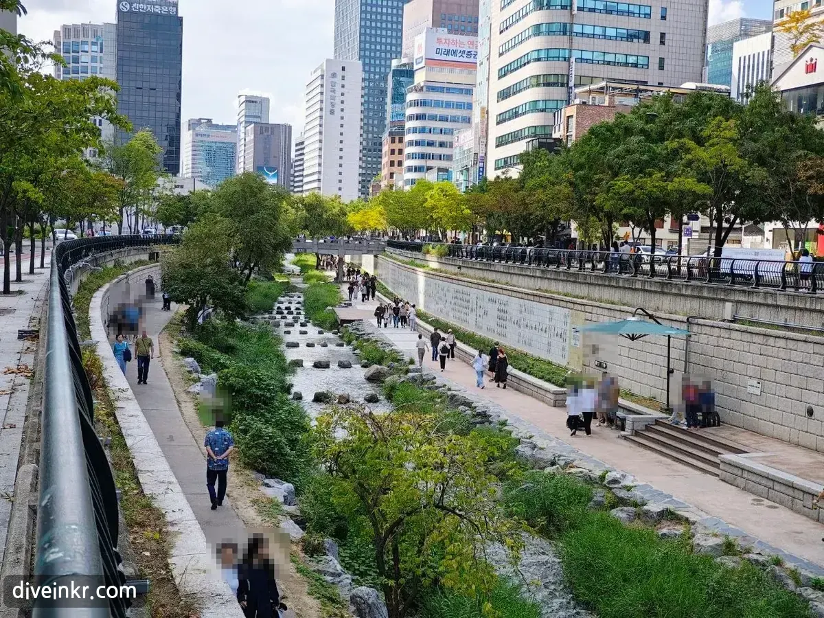 A wide view looking down Cheonggyecheon from street level: the stream with stepping stones running between two paved walkways lined with trees and stone walls, a small bridge in the distance and office towers on both sides (faces blurred)