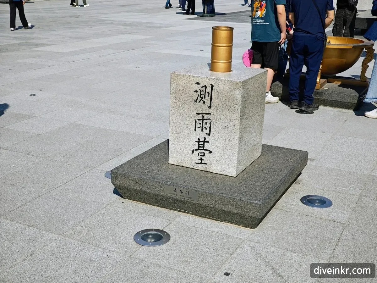 A copy of the Joseon rain gauge: a golden cylinder set into a granite block carved with the characters 測雨臺, on a dark stone base with a small plaque reading 측우기 Rain Gauge, in Gwanghwamun Square