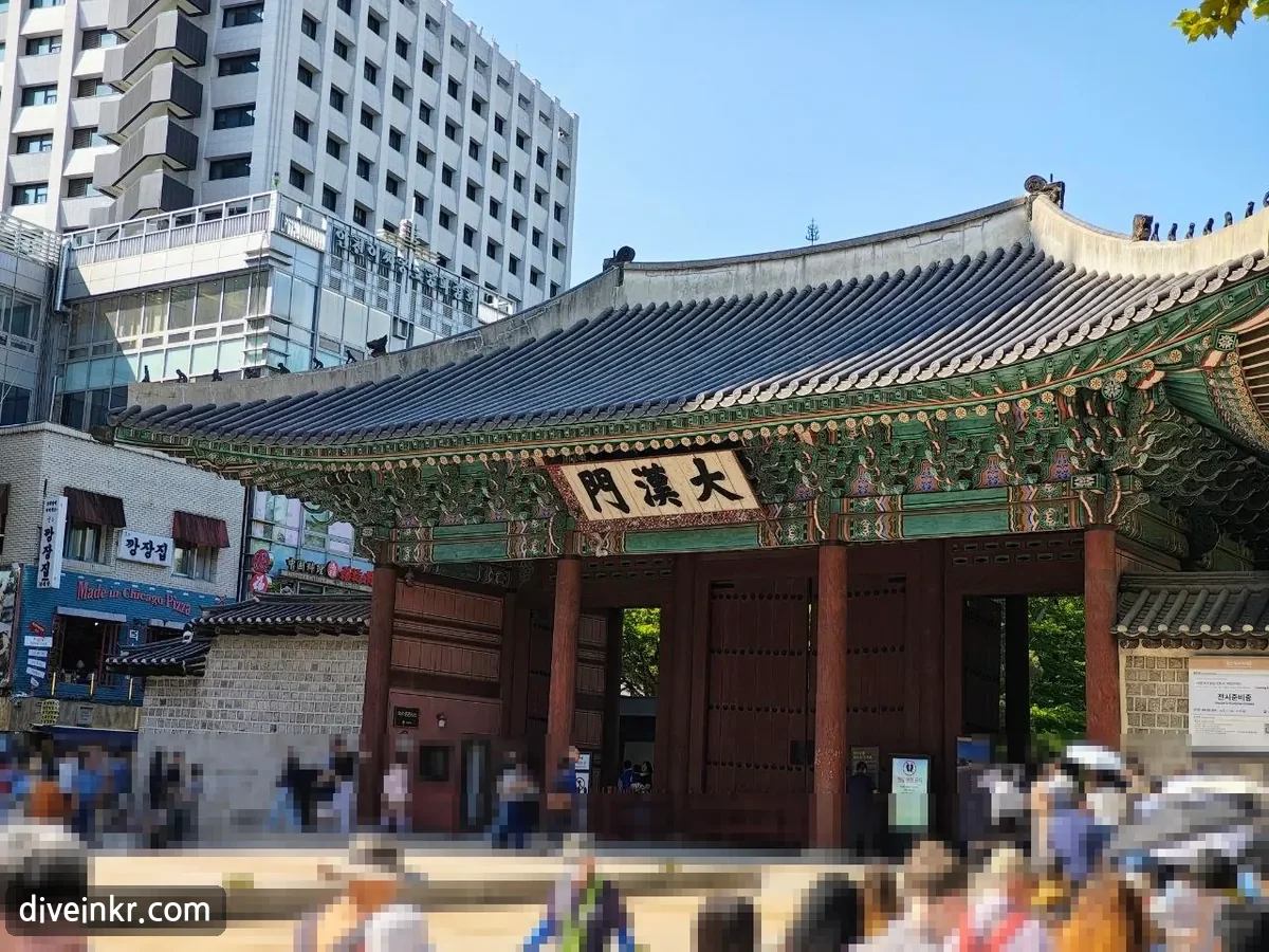 Daehanmun, the main gate of Deoksugung Palace: a wooden gate with a tiled roof, painted eaves, red pillars and doors and a name board reading 大漢門, standing between office buildings, with visitors in front (faces blurred)