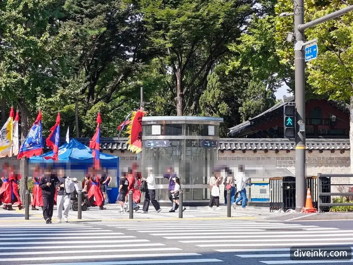 Guards in red Joseon-era uniforms carrying tall red, blue and yellow flags on the sidewalk beside Deoksugung's stone wall, next to a glass elevator entrance marked 시청 with the station numbers 132 and 201, seen across a crosswalk; a blue street sign on the right reads 소공로 Sogong-ro (faces blurred)