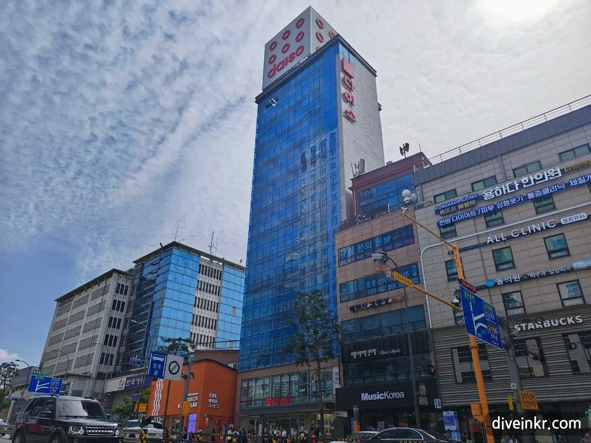 The Daiso Myeongdong Station store: a tall narrow blue-glass building with a red daiso logo and the vertical Korean sign λ€μ΄μ at the top, next to buildings with signs for a Korean medicine clinic, other clinics and a Starbucks, under a sky streaked with clouds