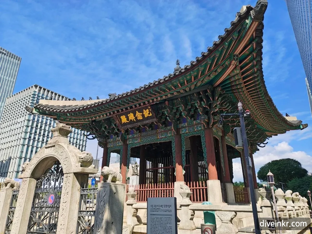 The pavilion for the monument to King Gojong's 40th year on the throne: a richly painted wooden pavilion with sharply upturned eaves and a name board reading 紀念碑殿, behind a stone gate topped with stone animals and an information panel, next to glass office towers