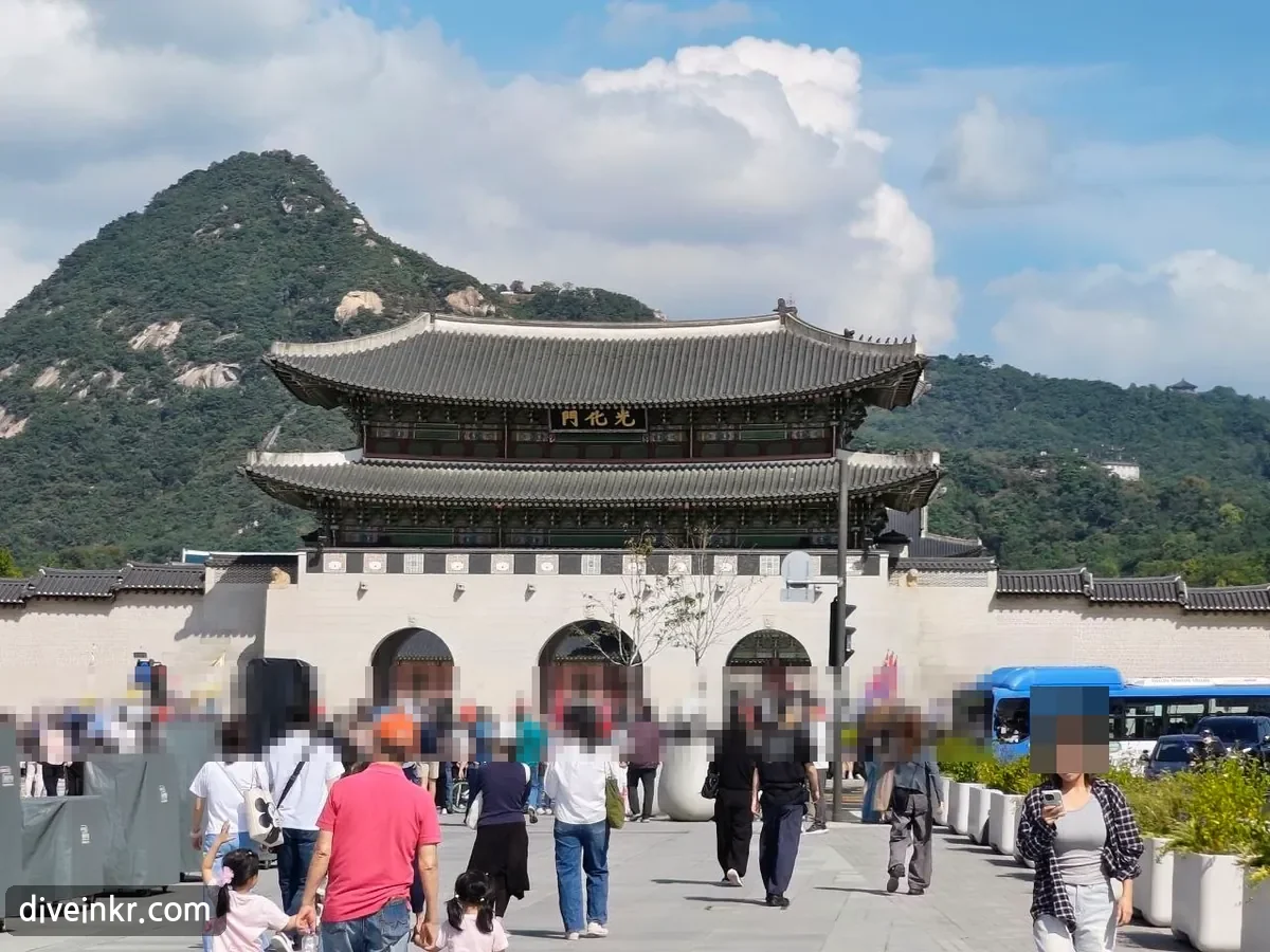 Gwanghwamun, the main gate of Gyeongbokgung Palace: a two-story wooden gate tower with a curved tiled roof and a black name board with gold characters 光化門, on a stone base with three arched openings, crowds of visitors in front and Bugaksan behind (faces blurred)