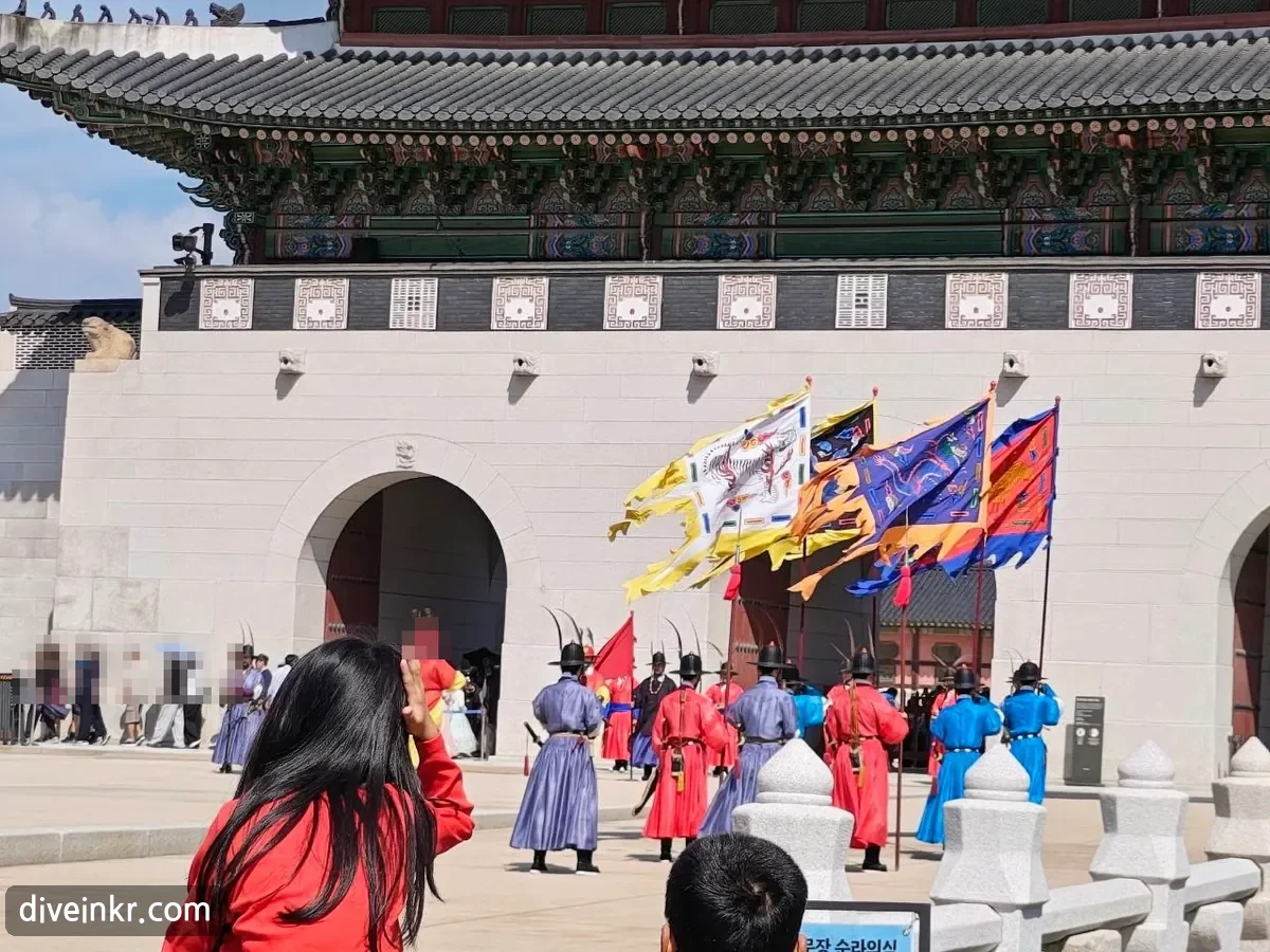 Close-up of the guards: men in red, blue and purple robes with black feathered hats and bows on their backs stand in formation in front of the stone base of Gwanghwamun, holding large painted flags, one showing a dragon, as spectators watch in the foreground; a blue sign at the bottom edge reads 순라의식 (faces blurred)