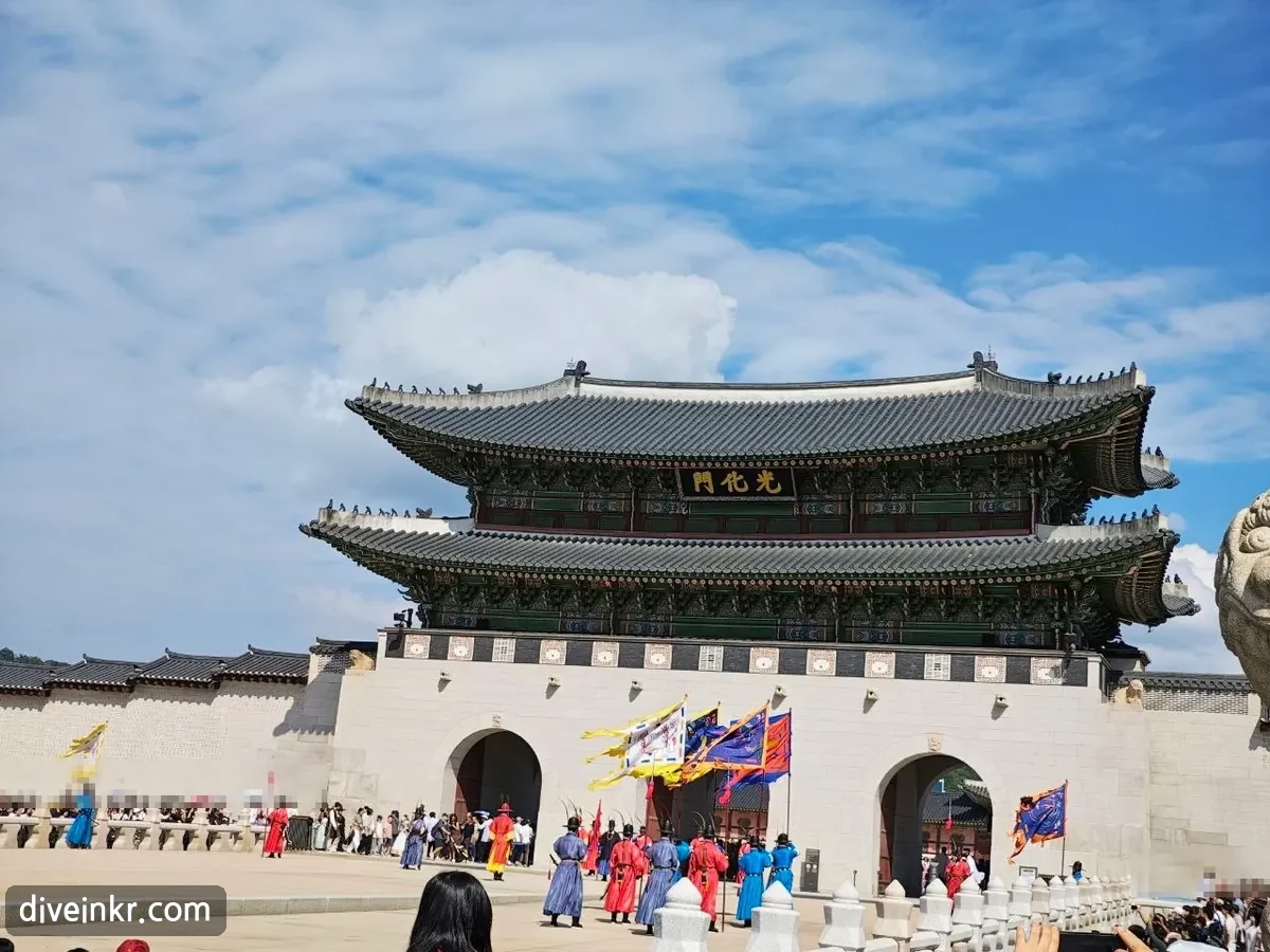 The royal guard ceremony in front of Gwanghwamun: guards in red, blue and purple Joseon-era uniforms stand in formation in front of the arched gateways, holding tall colorful flags, while crowds watch from behind stone railings (spectators' faces blurred)