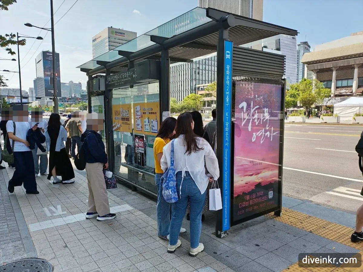 A bus shelter on the road beside Gwanghwamun Square with a blue sign reading 광화문역2번출구·KT광화문지사 and the English Gwanghwamun Station Exit 2 · KT (Gwanghwamun Bldg.), people waiting and a musical poster on the side panel (faces blurred)