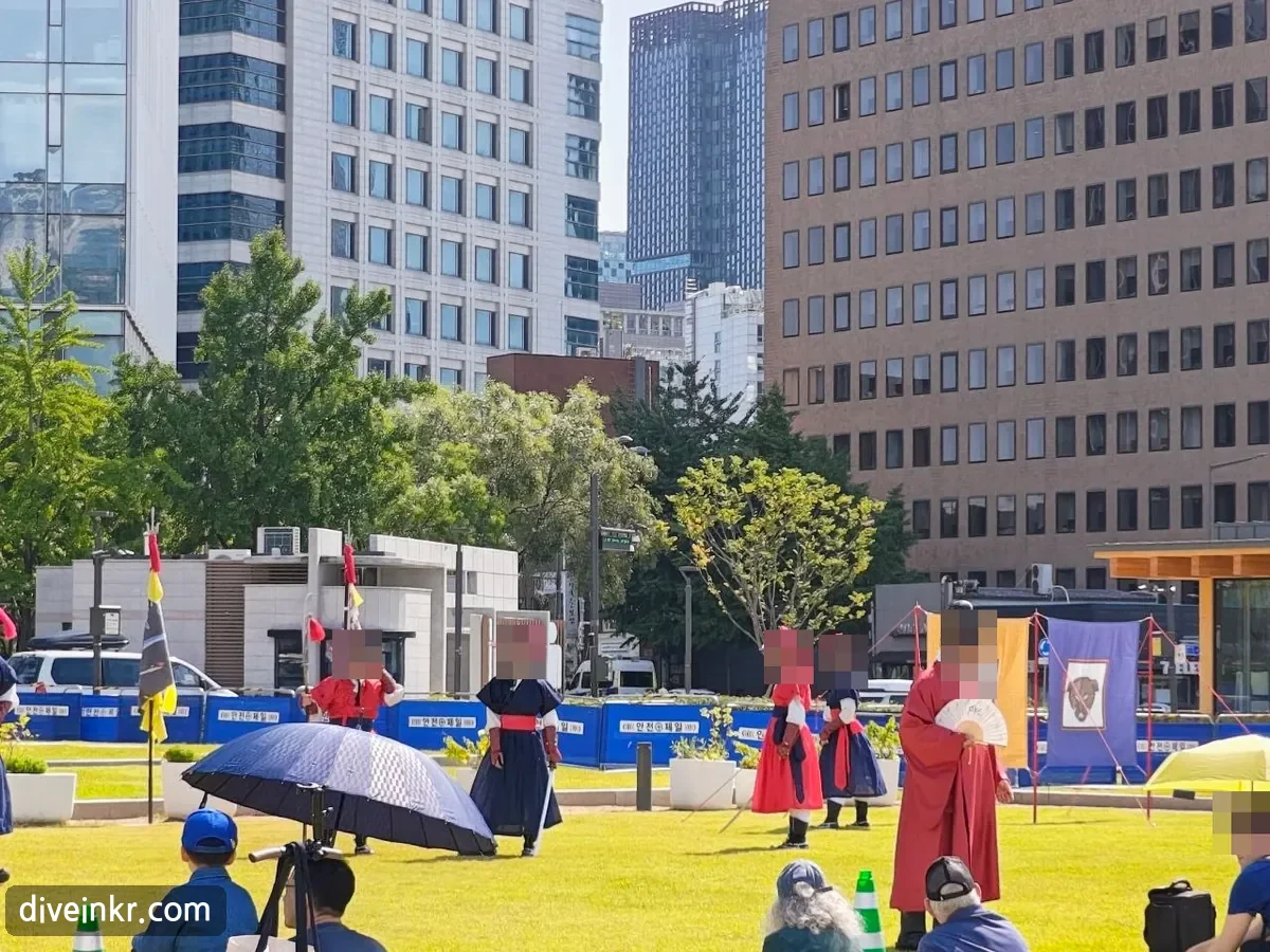 Performers in Joseon-era costumes — red and navy military uniforms with feathered hats, and a man in a red official's robe holding a fan — performing on the lawn beside Gwanghwamun Square, with banners and an audience seated on the grass (faces blurred)
