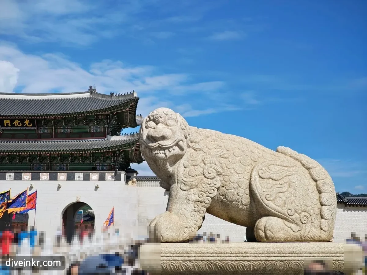 A large stone haechi statue with curly carved mane, bulging eyes and a toothy grin, crouching on a stone plinth in front of Gwanghwamun gate, with guards in colorful uniforms and flags by the gate and visitors below (faces blurred)
