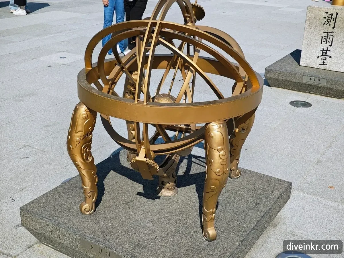 A golden copy of a Joseon armillary sphere: interlocking rings with a small globe in the center, set on four curved legs decorated with cloud patterns, on a stone base in Gwanghwamun Square