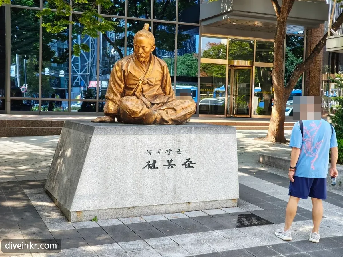 A bronze statue of Jeon Bong-jun seated cross-legged in traditional clothes on a granite base engraved 녹두장군 전봉준, outside a glass building near Jonggak Station, with a passerby standing beside it (face blurred)