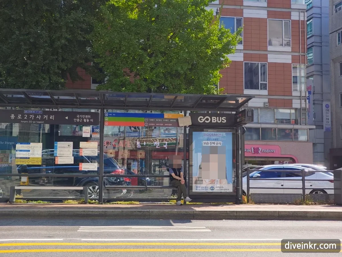 A bus shelter at Jongno 2-ga in Seoul: a dark sign reads 종로2가사거리 with the English Jongno 2(i)-ga Sageori and a small stop ID box, colored route bands and timetables on the glass, and a man waiting on the bench (face blurred)