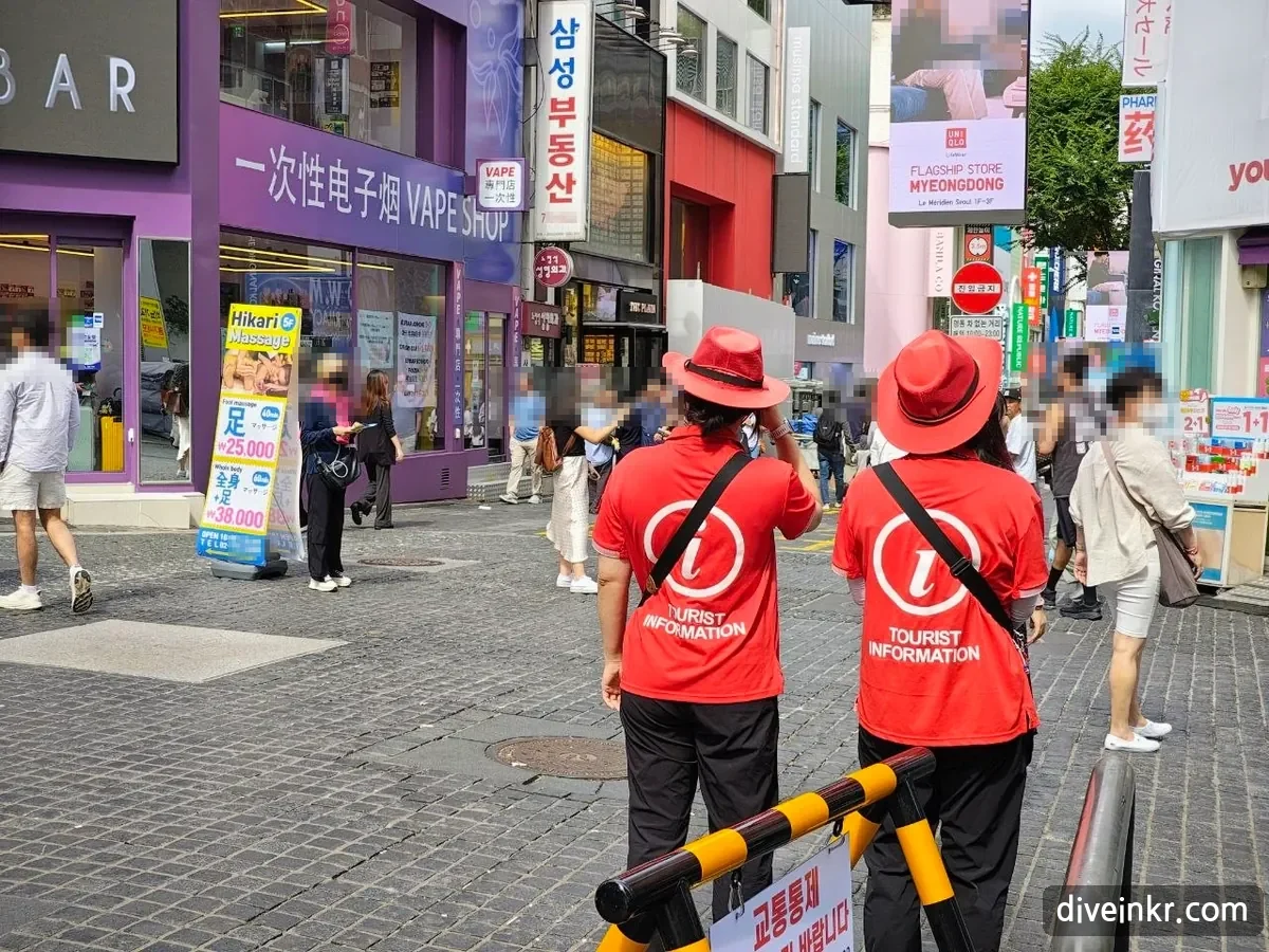 Two roaming tourist guides seen from behind in Myeongdong, wearing wide-brimmed red hats and red shirts printed with a white i and TOURIST INFORMATION, standing behind a yellow-and-black barrier with a sign reading κ΅ν΅ν΅μ and looking out over a busy shopping street with a no-entry sign in the distance (bystanders' faces blurred)