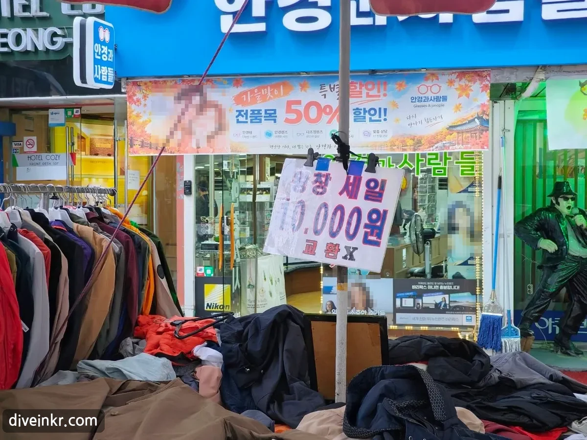 A handwritten sign clipped to a parasol pole above a pile of jackets, reading 왕창 세일 10.000원 교환 X. Behind it is an optician's shop with a blue sign and an autumn sale banner reading 가을맞이 특별할인! 전품목 50% 할인! (faces blurred)