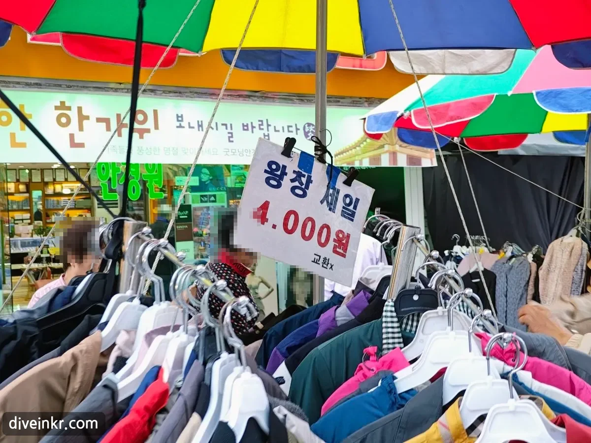 A handwritten sign reading 왕창 세일 4.000원 교환x clipped above racks of jackets under colorful parasols, with a shop sign behind reading 한가위 보내시길 바랍니다 and a green optician's sign (faces blurred)