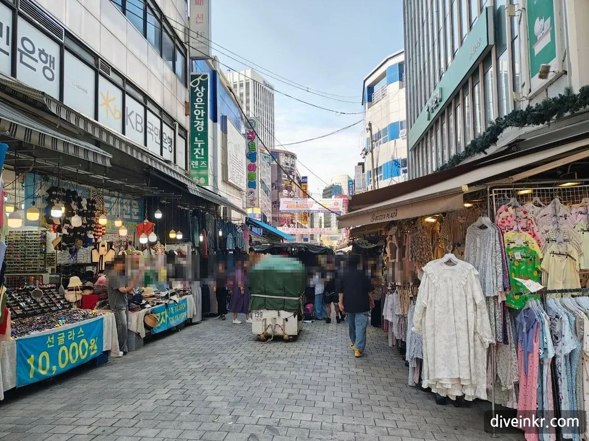 A street in Namdaemun Market lined with stalls selling sunglasses and hats, with a blue banner reading 선글라스 10,000원, racks of dresses on the right, a covered cart in the middle of the lane, a KB Kookmin Bank sign on the building at left and shoppers walking (faces blurred)