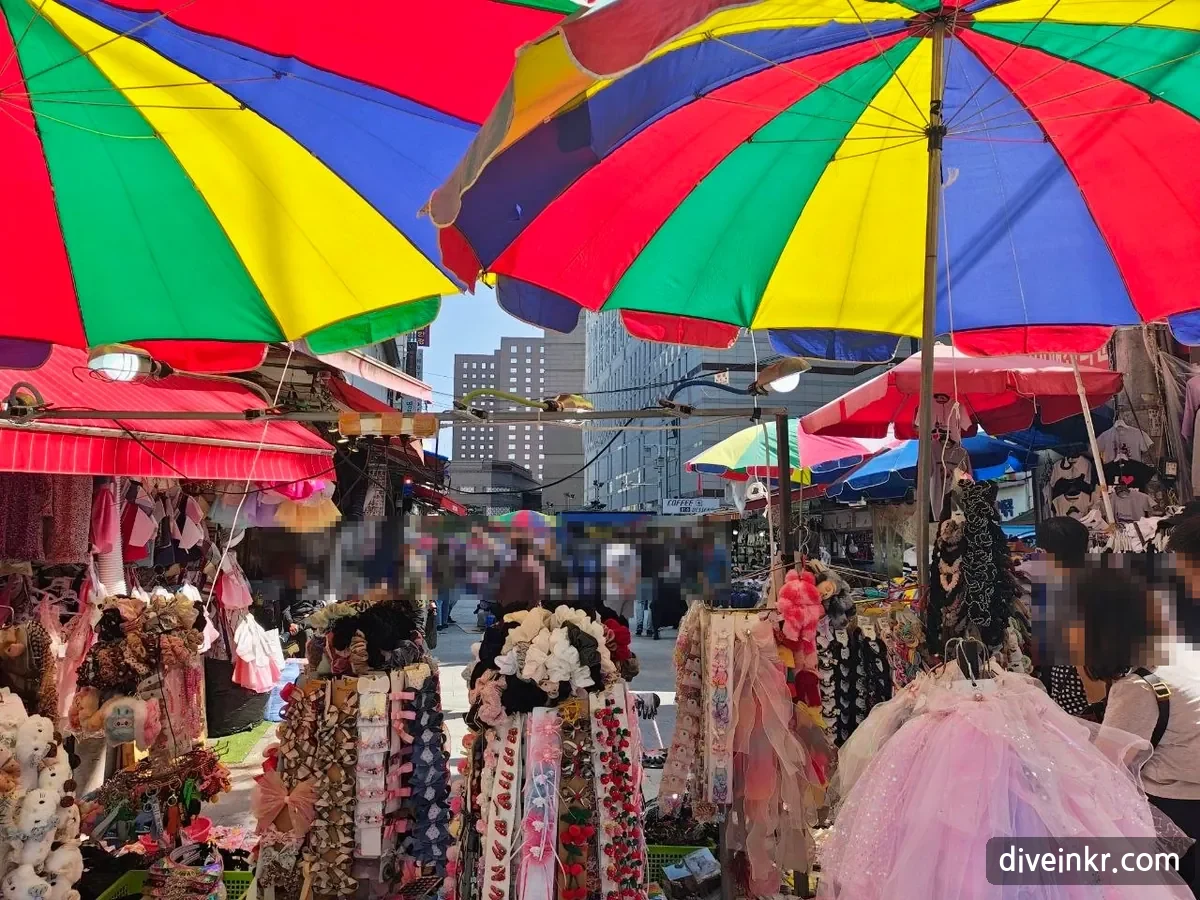 Stalls under large rainbow-striped parasols in Namdaemun Market, crowded with racks of hair clips, scrunchies, children's hair accessories and a pink tulle dress, with shoppers in the lane behind (faces blurred)
