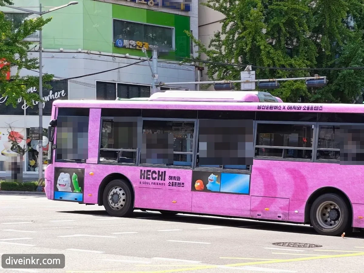 A pink λ¨μ°μνλ²μ€ (Namsan loop bus) wrapped with the ν΄μΉμ μμΈνλ μ¦ characters, driving past shops (passengers' faces blurred)