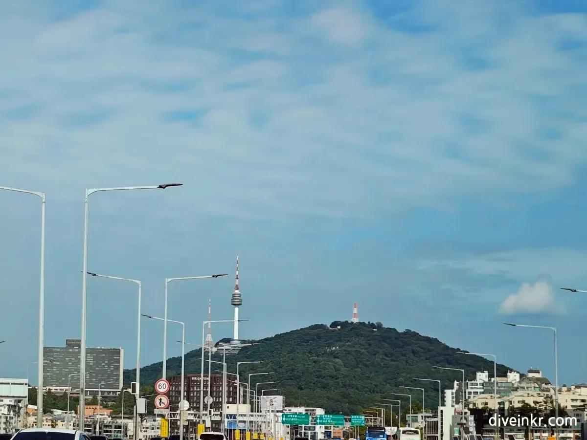N Seoul Tower standing on the green hill of Namsan under a blue sky with light clouds, seen from a road in Seoul lined with street lamps and road signs