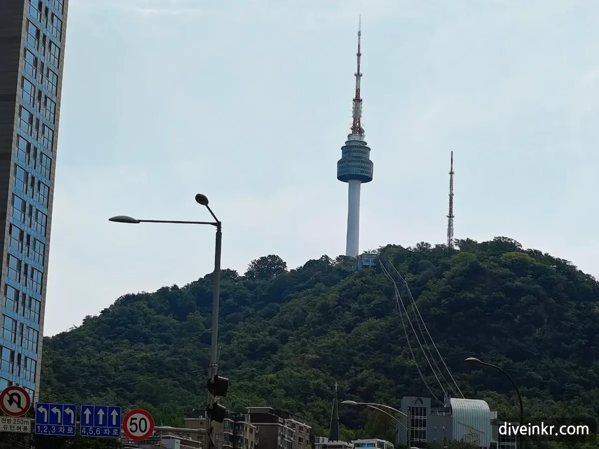N Seoul Tower on the wooded summit of Namsan seen from below, with a second transmission mast to the right and cables running down the hillside, above a no-U-turn sign with a plate reading μ λ°© 250m μ ν΄νμ©, blue lane signs reading 1,2,3 μ°¨λ‘ and 4,5,6 μ°¨λ‘, and a 50 km/h speed-limit sign