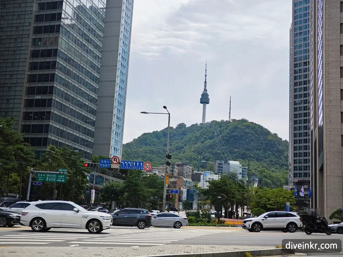 A wide downtown intersection with cars crossing, office towers on both sides, and Namsan with N Seoul Tower rising in the gap between the buildings; a green direction sign at left, under a smaller sign reading ννμ¬κ±°λ¦¬, points left to λͺ
λμ and right to μμΈμ (license plates blurred)
