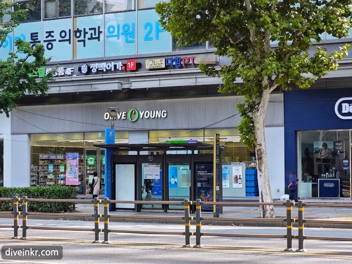 An Olive Young store on a Jongno street, with the OLIVE YOUNG sign above glass windows, a bus shelter in front, and signs on the upper floors reading 통증의학과 의원 2F, 정백어가 1F and a PT studio on the 4th floor (faces and a phone number blurred)
