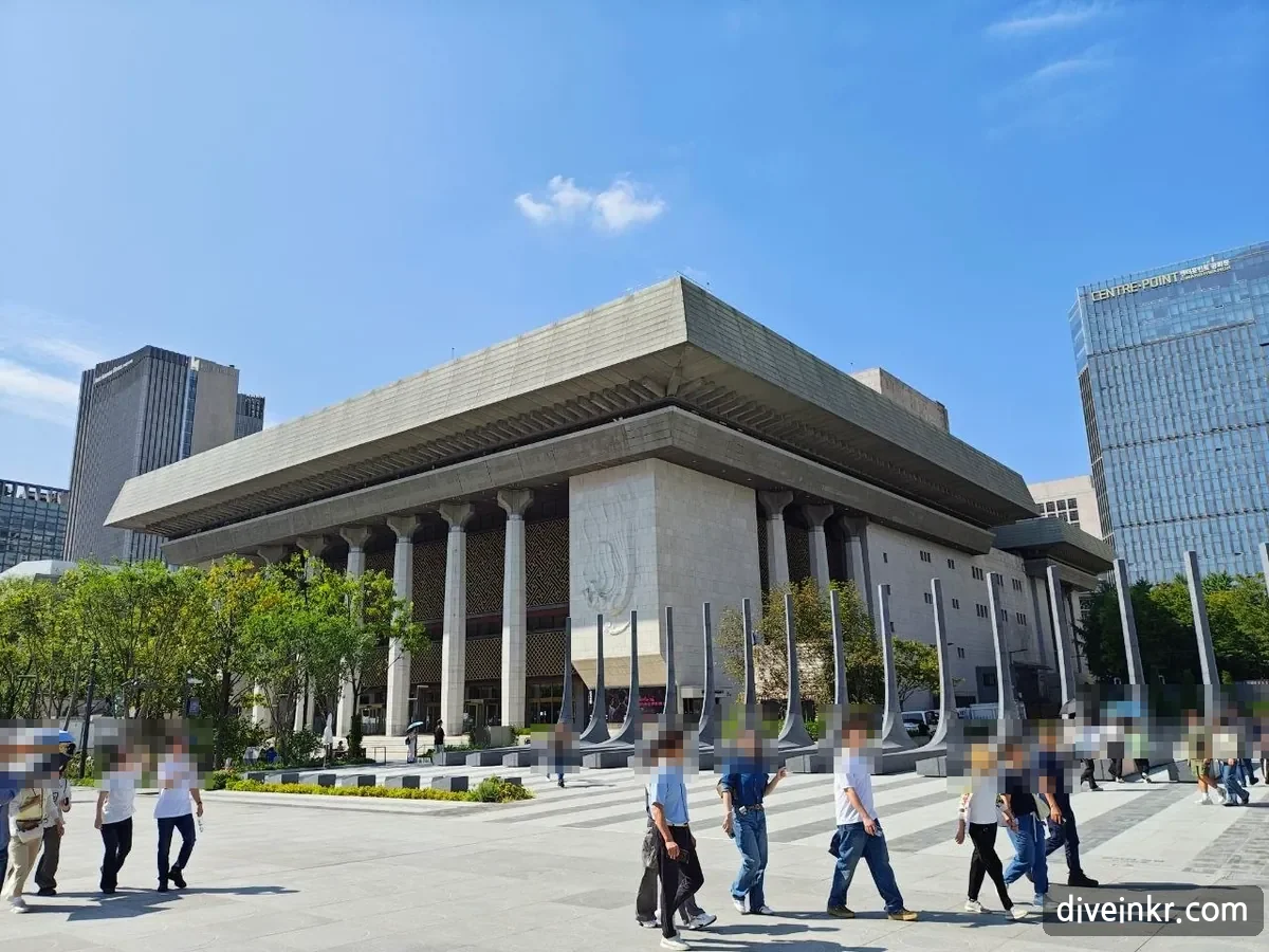 The Sejong Center for the Performing Arts on the west side of Gwanghwamun Square: a large stone building with a wide overhanging roof and tall columns, with visitors walking across the plaza in front (faces blurred)