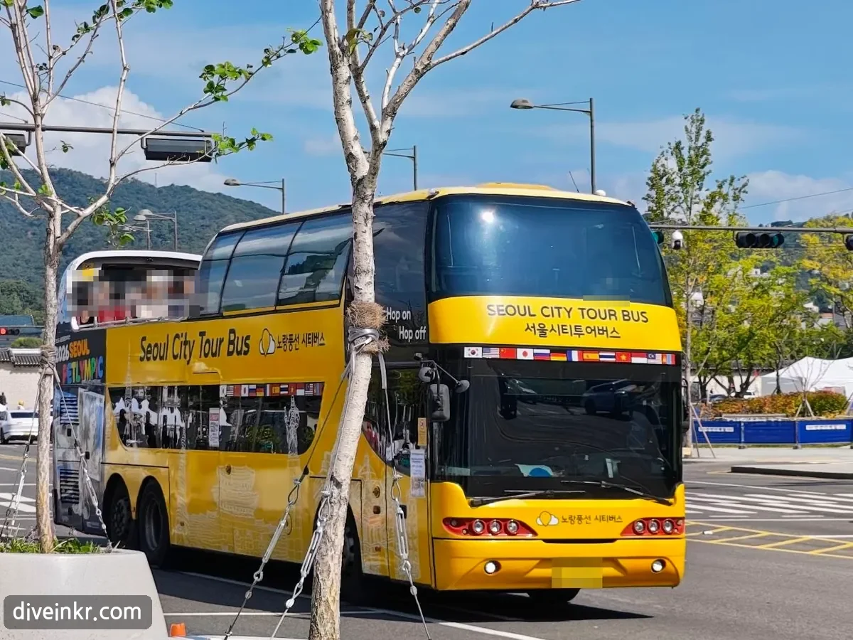 A yellow double-decker tour bus near Gwanghwamun with SEOUL CITY TOUR BUS and 서울시티투어버스 above the windshield, a row of national flags, Hop on Hop off on the side window and the 노랑풍선 시티버스 name on the front (license plate and passengers blurred)