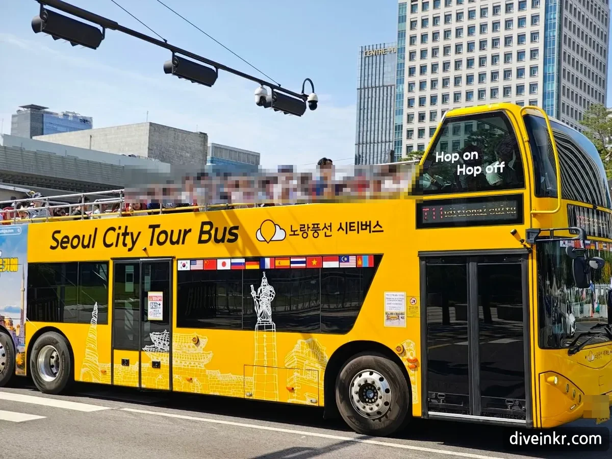 A yellow open-top double-decker bus reading Seoul City Tour Bus and 노랑풍선 시티버스, with Hop on Hop off on the front window, flags of many countries along the side and passengers on the upper deck (faces blurred), passing office buildings near Gwanghwamun