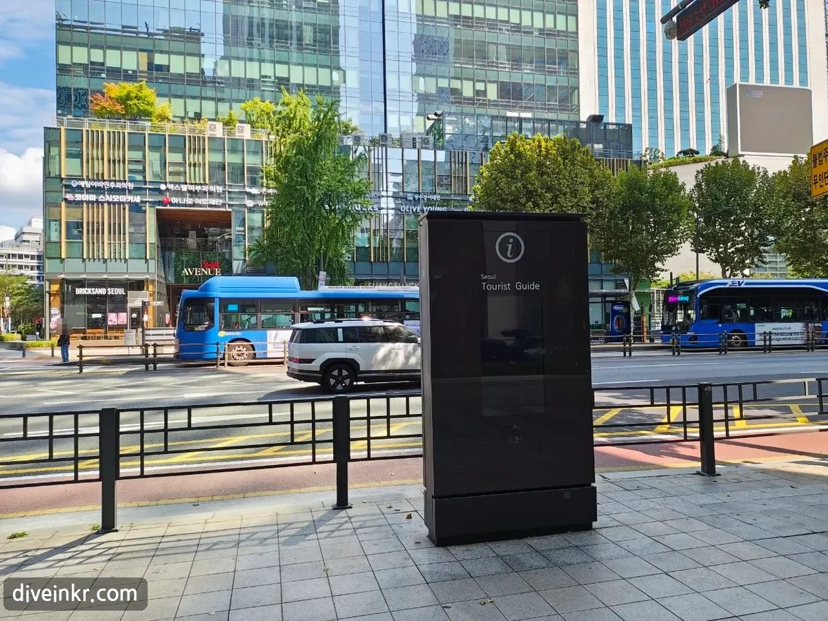 A tall black street kiosk with an i symbol and the words Seoul Tourist Guide on a city sidewalk in central Seoul, with blue buses and glass office buildings across the road