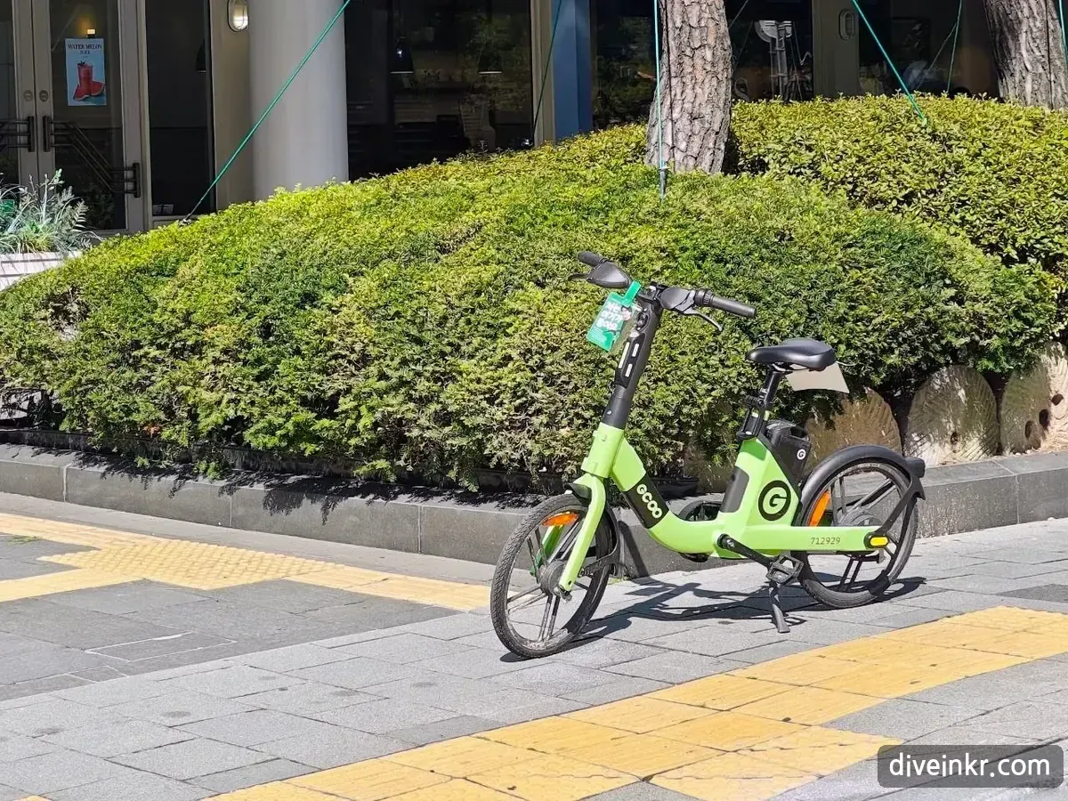 A green shared electric bicycle from a private company parked on a paved sidewalk next to a trimmed hedge in Seoul, with a yellow tactile paving strip in the foreground