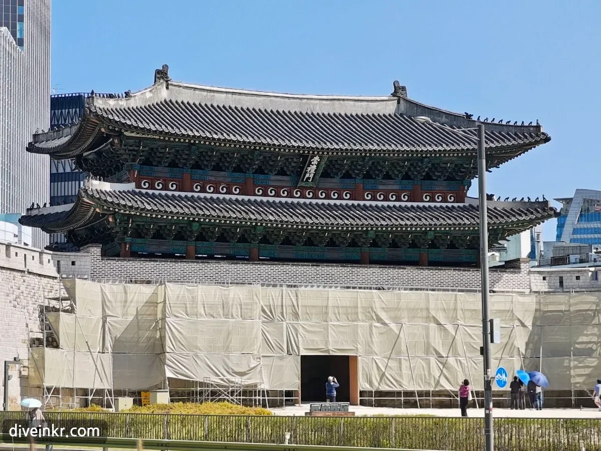 A close view of Sungnyemun's two-tiered tiled roof and painted eaves, with a vertical name board reading 崇禮門 hanging between the two roofs; the stone base below is wrapped in beige construction screens with an opening left at the central passage, and a few visitors stand in front
