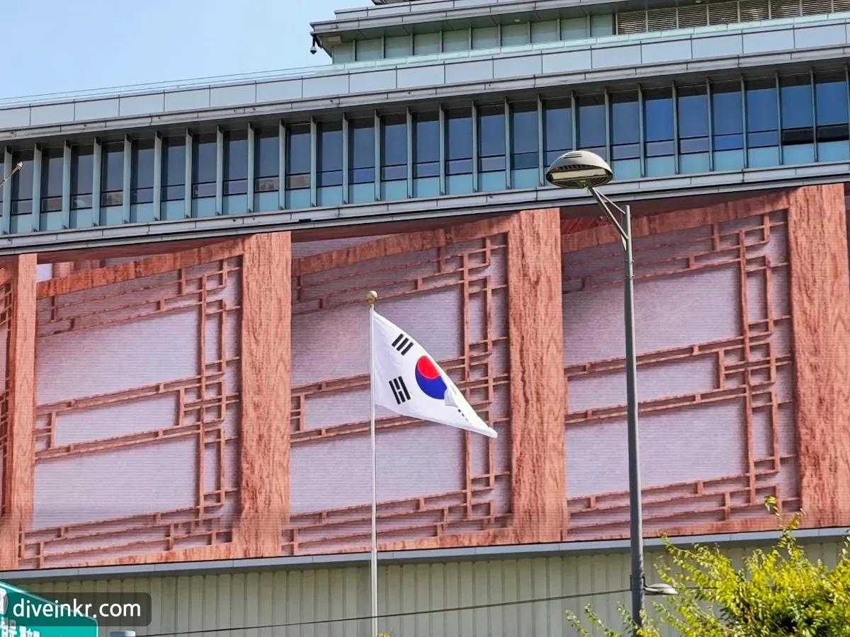 The Korean national flag, Taegeukgi, flying from a pole in front of a building near the Seoul Museum of History, whose facade shows a large image of a traditional wooden lattice window
