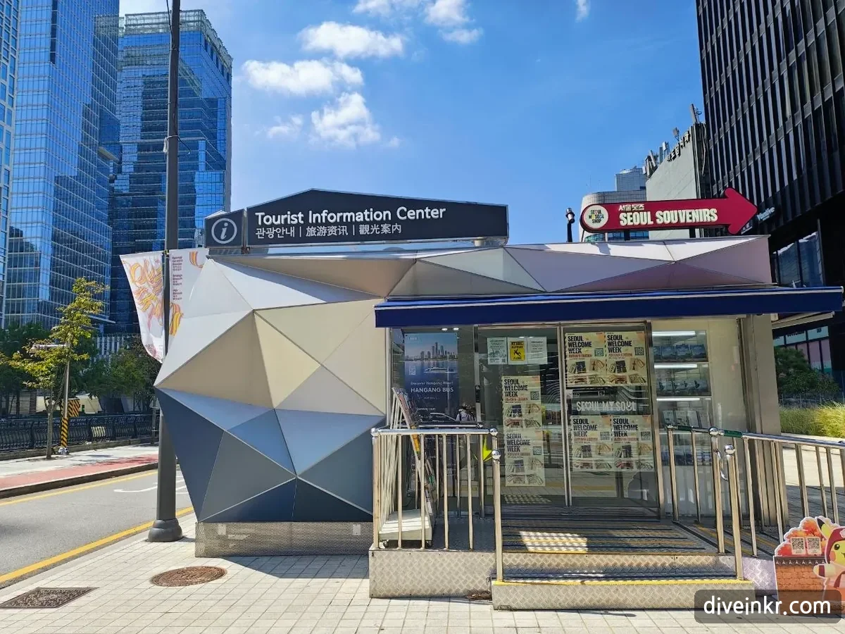 A small Tourist Information Center booth with an angular silver facade and a sign reading κ΄κ΄μλ΄ in Korean, Chinese and Japanese, with Seoul Welcome Week posters in the windows and a red arrow sign reading SEOUL SOUVENIRS beside it, on a sidewalk beside Cheonggyecheon stream